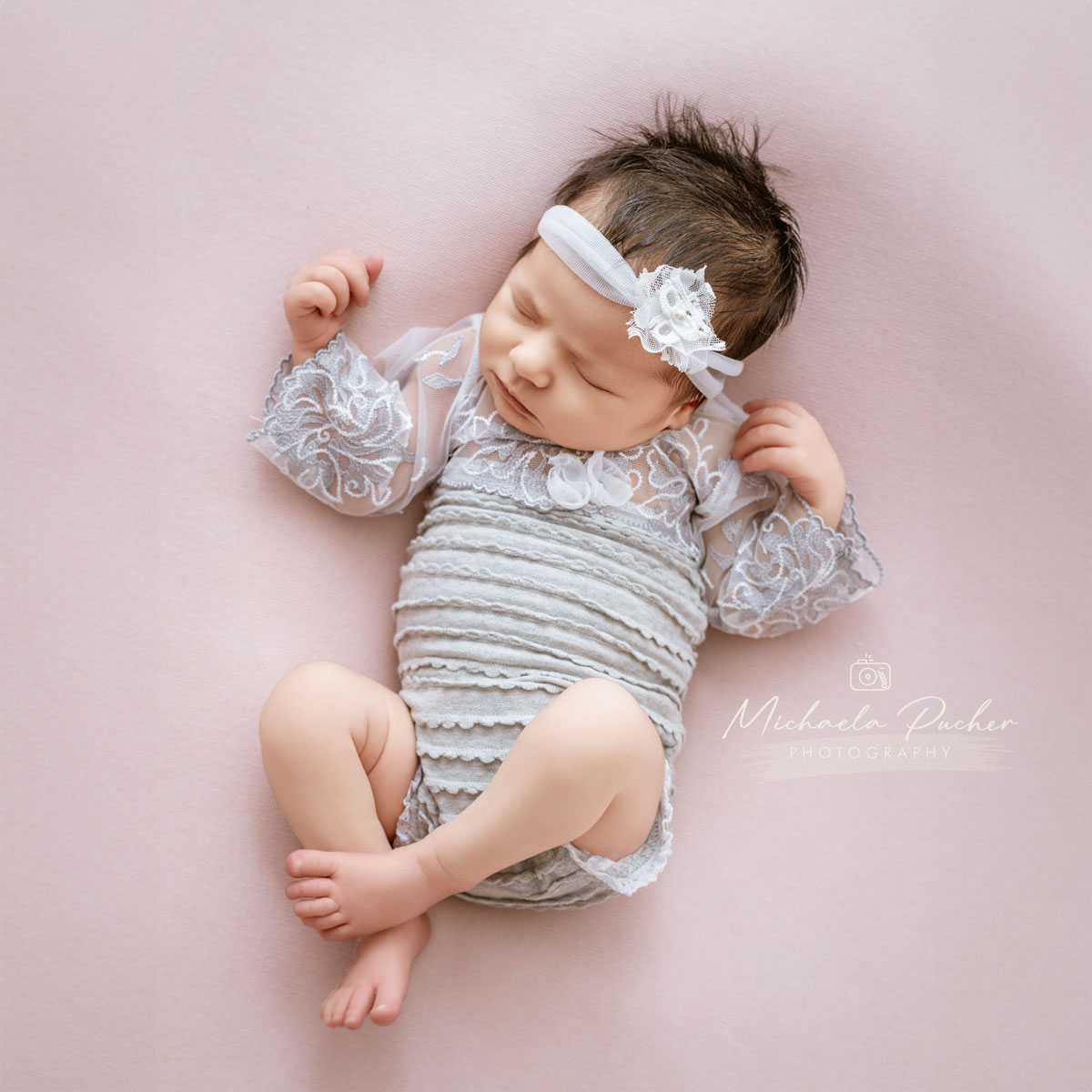 Neugeborenes Mädchen schläft entspannt auf einer zart rosafarbenen Unterlage, trägt ein hellgraues Spitzen-Outfit und ein passendes Haarband.